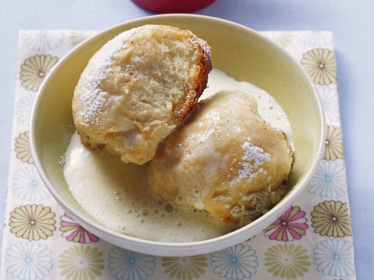 Zwei Dampfnudeln mit Vanillesoße in einer hohen, runden Schale aus Porzellan / Untergrund quadratische papierserviette auf glattem Boden