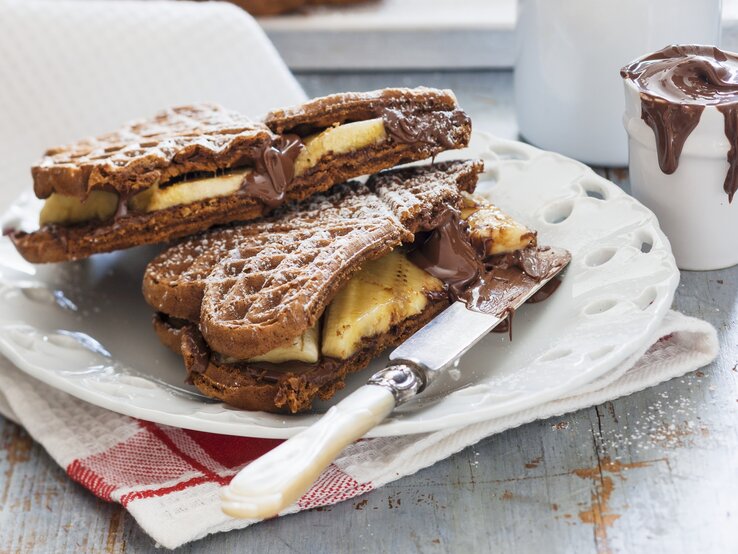 Schoko-Waffeln mit Banane auf einem weißen Teller auf einem Geschirrtuch. Auf dem Teller liegt ein Messer und hinten auf dem Bild ein Glas mit Schoko-Creme.