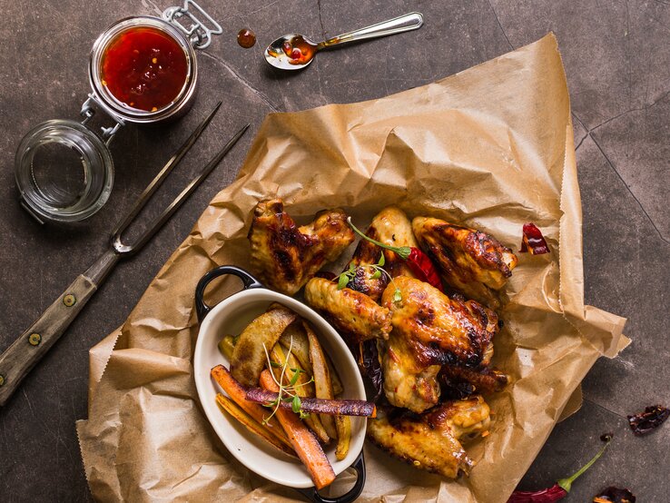 Hähnchenflügel-Chilimarinade Knusprige Hähnchenflügel mit geschmortem Gemüse auf Backpapier. Alles direkt von oben auf einem grauen Tisch fotografiert.