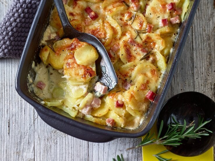 Kartoffelauflauf mit Schinken in ofenfester Form / großer Löffel aus Plastik / Untergrund Holz / Draufsicht