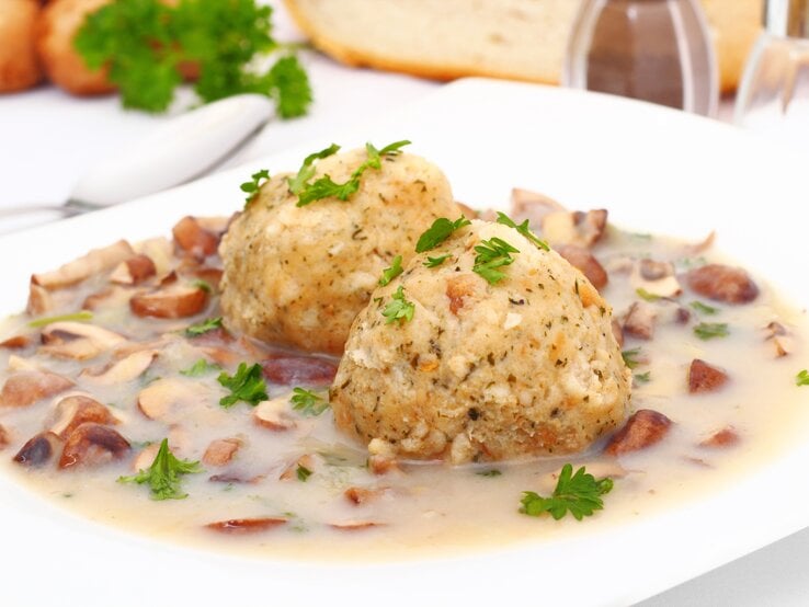 Auf einem weißen Teller liegen zwei Semmelknödel, garniert mit frischer Petersilie, umgeben von einem cremigen Ragout mit kleinen Pilzstücken. Im Hintergrund sind Brot, Kräuter und Geschirr zu erkennen.