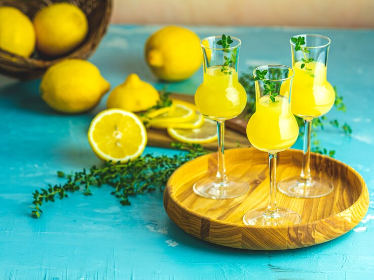 Mehrere Gläschen gefüllt mit Limoncello auf einem Holzbrett. Dahinter liegen mehrere Zitronen auf blauem Untergrund. 