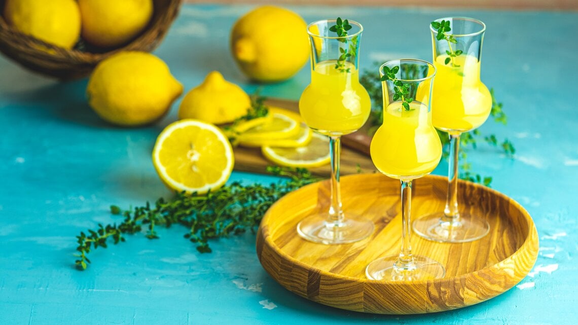 Mehrere Gläschen gefüllt mit Limoncello auf einem Holzbrett. Dahinter liegen mehrere Zitronen auf blauem Untergrund. 