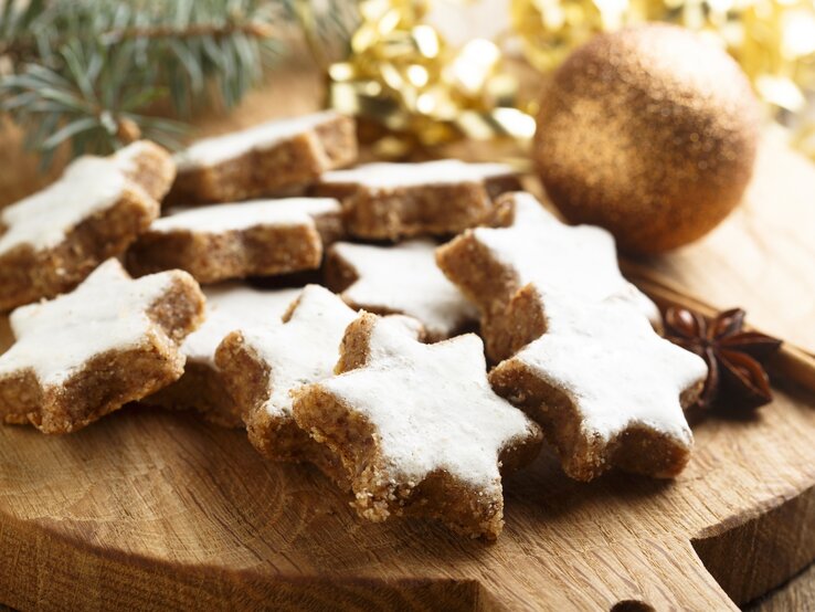 Mehrere frisch gebackene Zimtsterne auf einem Holzbrett. Daneben weihnachtliche Dekorationselemente wie Tannenzweige und goldenes Lametta. 