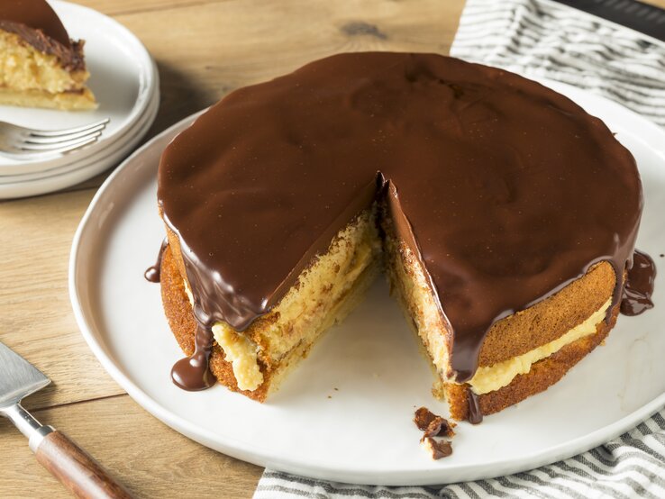 Ein Bild von einem Boston Cream Pie auf einem weißen Teller, mit einem Stück herausgeschnitten, zeigt die Schichten aus fluffigem Kuchen, cremiger Vanillefüllung und glänzender Schokoladenglasur, serviert auf einem Holztisch mit einem gestreiften Küchentuch im Hintergrund.