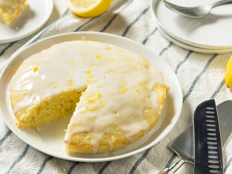 Runder, angeschnittener Zitronenkuchen mit heller Glasur auf einem weißen Teller. 