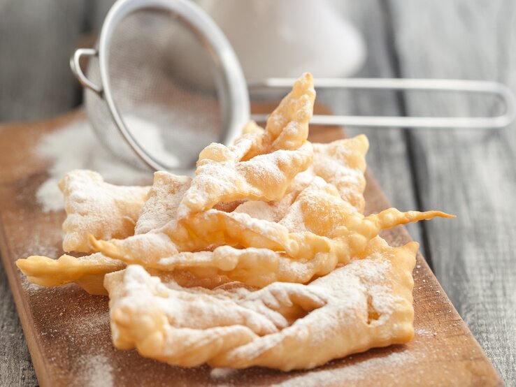 Ein Stapel frisch gebackener Faworki (auch bekannt als Engelshaar oder Krapfen), ein traditionelles polnisches Gebäck, das zu Karneval und Fasching serviert wird. Die Faworki sind goldbraun und mit Puderzucker bestäubt, der durch ein kleines Metallsieb gestreut wurde, das im Hintergrund unscharf zu sehen ist.