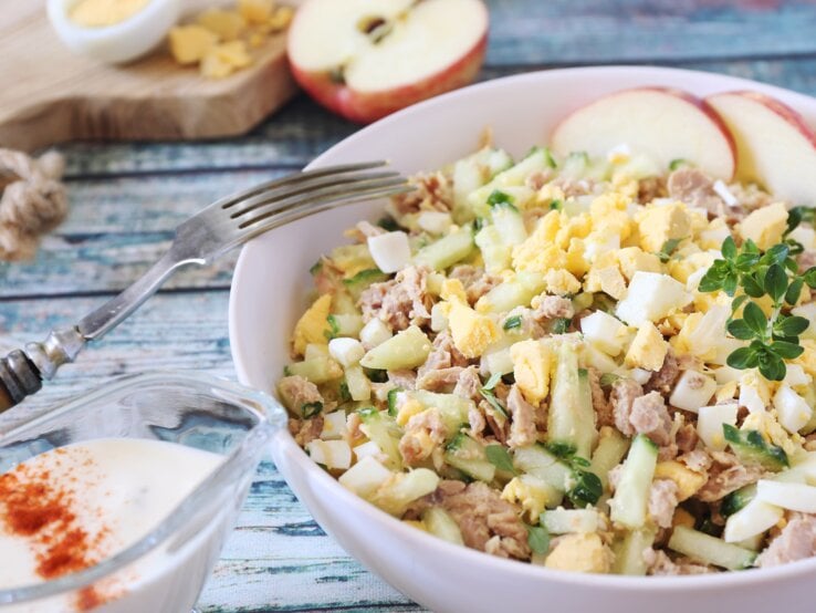 Cremiger Salat aus Ei, Thunfisch und Gurken, mit Apfelscheiben und Kräutern garniert, serviert auf einem Holztisch.