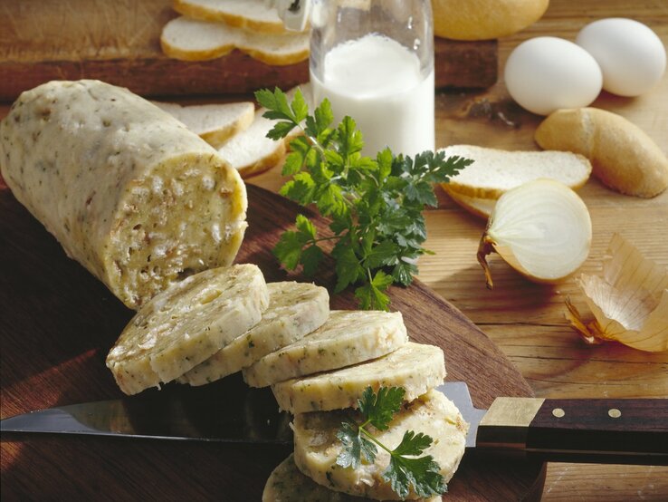 Auf einem rustikalen Holzbrett liegen aufgeschnittene Böhmische Knödel, dekoriert mit frischer Petersilie. Daneben stehen Brot, Eier und eine halbierte Zwiebel.