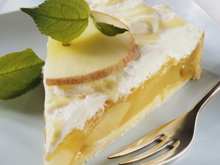 Nahaufnahme eines Stücks Eierlikör-Apfel-Torte mit Vanillecreme, frischen Apfelscheiben und Minzblättern auf einem weißen Teller, daneben eine Kuchengabel