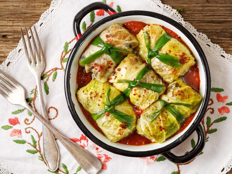 Frisch gemachte vegetarische Kohlrouladen in einem Topf / jede ist mit einem breitem band verschnürt / Untergrund besticktes Tuch auf Holz / Zwei gabeln / Draufsicht