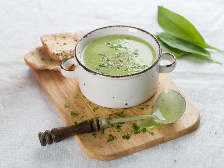 Ein Topf mit cremiger Bärlauchsuppe, bestreut mit Kräutern, neben einem Stück Brot auf einem Holzbrett, mit einem alten Löffel und frischen Bärlauchblättern daneben.