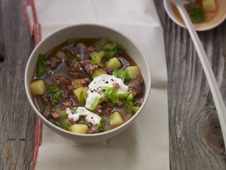 Eine weiße Schale mit dem deftigem Eintopf auf einem rustikalen Holztisch. Alles von schräg oben fotografiert.