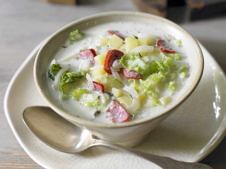 In einer beigefarbenen Suppenschale steht eine cremige Wirsingsuppe mit Wurst. In der hellen Brühe schwimmen Stücke von grünem Wirsing, gelben Kartoffelwürfeln und roten Wurstscheiben. Die Suppe steht auf einem Teller, daneben liegt ein Löffel. Im Hintergrund ist ein unscharfer Holztisch zu erkennen, der der Szene eine warme, gemütliche Atmosphäre verleiht.