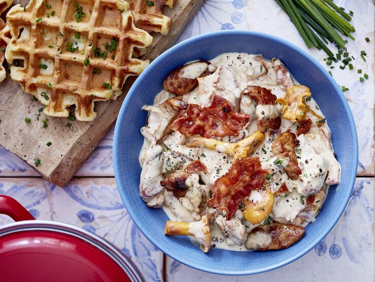 Pfifferlinge und Bacon machen dieses Rezept für Filet-Gulasch erst richtig lecker! Ein Teller mit Rahmgeschnetzeltem aus Pilzen und Speck, serviert in einer blauen Schale, daneben frische herzhafte Waffeln mit Schnittlauch.