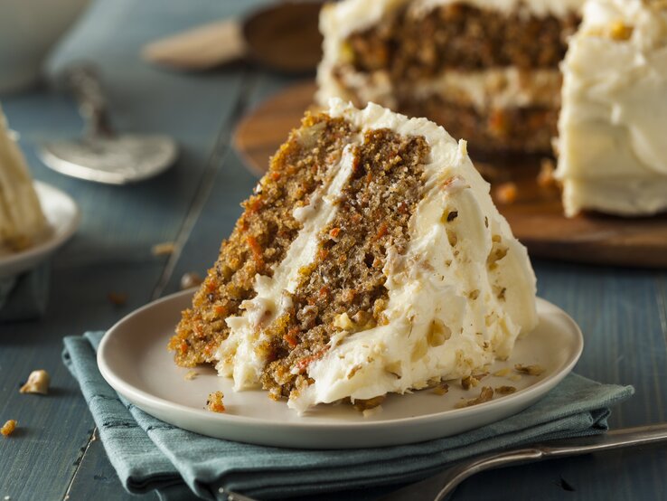 Ein Stück Karotten-Nuss-Kuchen mit Frischkäse auf  weißem Teller auf blauem Untergrund.