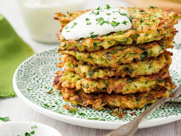 Mehrere der Spitzkohl-Röstis auf einem weiß-grünen Teller gestapelt. Oben dann noch ein Klecks Joghurt und frische Kräuter. Alles von der Seite auf einem hellen Holztisch fotografiert. 