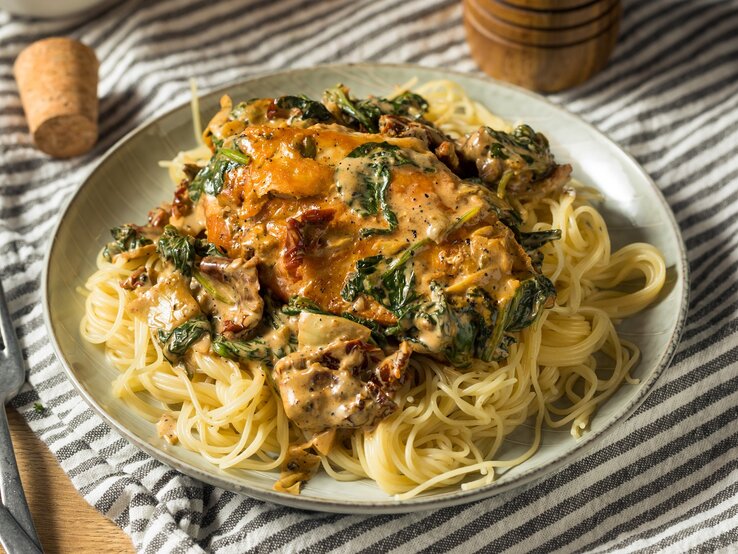 Spaghetti mit Paprika-Sahne-Hähnchen und Spinat Ein grauer Teller mit dem Pastagericht auf einer weiß-schwarz-karierten Tischdecke. Alles auf einem Holztisch mit Salz- und Pfefferstreuer aus Holz im Hintergrund.