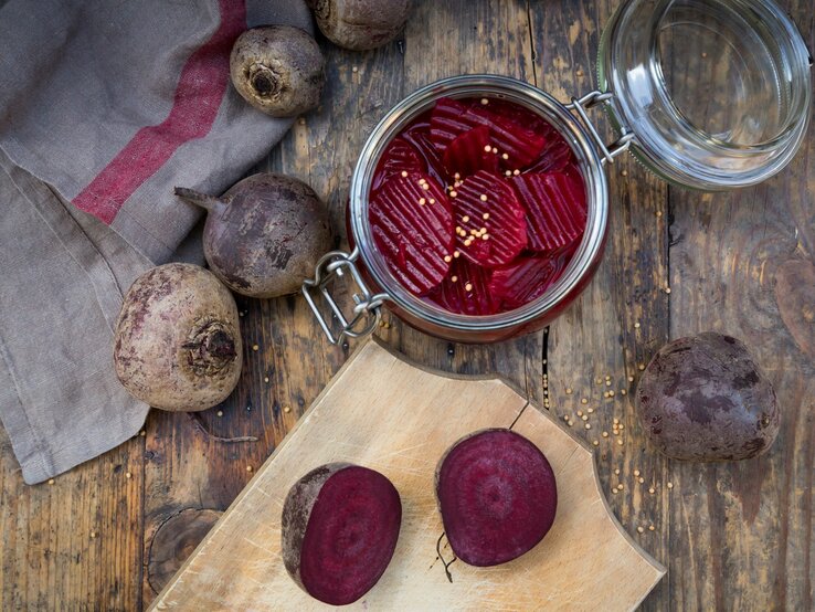 Rote Bete in einem Glas mit geöffnetem Decken. Im Bild eine aufgeschnittene Rote Bete auf Holzbrett. Weitere im Bild verteilt. Arrangiert auf Holztisch. Vogelperspektive.