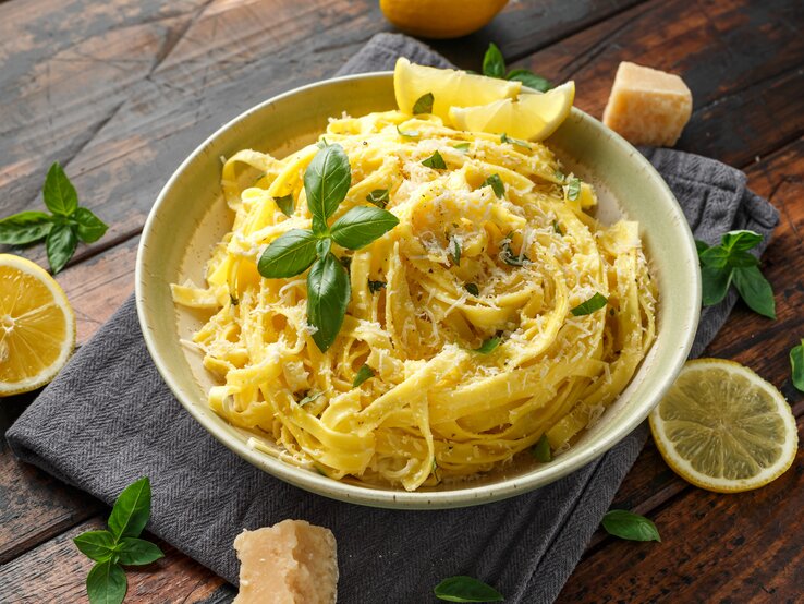 Ein Teller mit Linguine al Limone in einer cremigen Zitronensauce, garniert mit frisch geriebenem Parmesan und Basilikumblättern. Neben dem Teller liegen Zitronenscheiben, frische Basilikumblätter und ein Stück Parmesan auf einem dunklen Holztisch. | © GettyImages.de/DronG