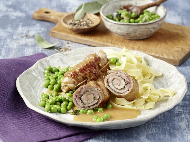 Saltimbocca Kalbsrouladen Ein Teller Saltimbocca Kalbsrouladen, dazu breite Bandnudeln und grüne Erbsen, angerichtet auf einer hellen Keramikplatte.