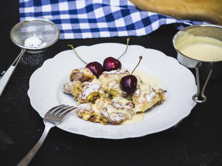 Kaiserschmarrn mit Kirschen und Vanillesauce aus einem runden Teller / Sieb mit Puderzucker / Draufsicht, schräg