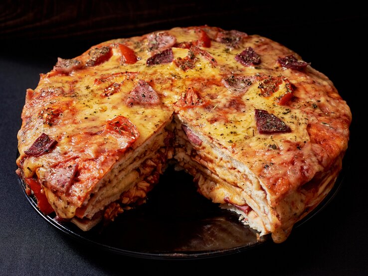 Gestapelter Pizzakuchen mit Käse, Schinken und Salami in dunkler Kulisse. 