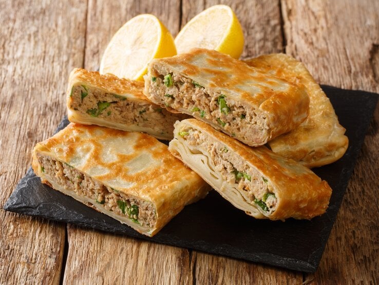 Eckig gefaltete Pfannkuchentaschen mit saftiger Hackfleisch-Füllung auf Schieferplatte. Diese steht auf einem rustikalen Holztisch. 