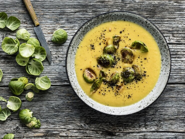 Draufsicht: Hölzerner Untergrund auf der ein grauer Steingutteller mit Steckrübensuppe und Rosenkohl-Topping steht. Links daneben noch Schalen frischer Rosenkohlröschen und ein scharfes Messer mit Holzgriff.
