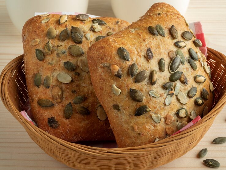 Selbst gemachte Kürbiskernbrötchen in Brotkorb auf Holztisch. Im Bild zwei weiße Tassen und einige Kürbiskerne. Frontalansicht.