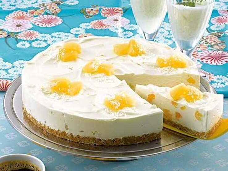 Pina-Colada-Torte mit Ananas-Stücken / Angeschnitten / 1 Stück Torte auf Tortenheber /  Zwei gefüllte Cocktailgläser / Untergrund Tisch / Draufsicht schäg