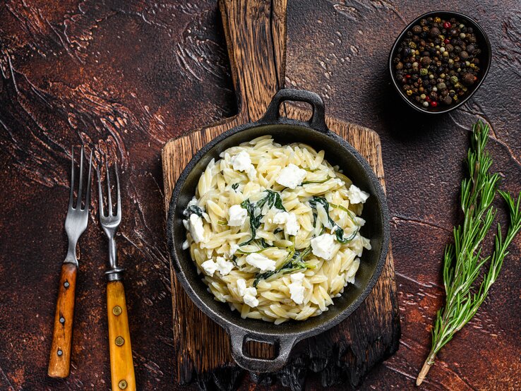 Gusseiserne Pfanne gefüllt mit Nudelreis, Spinat und Käse in rustikalem Ambiente mit mediterraner Dekoration.