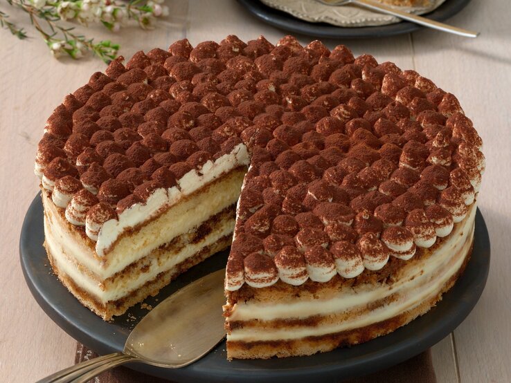 Die angeschnittene Kaffee-Schoko-Torte auf einem dunklen Tortenständer und beigem Holztisch. Alles von schräg oben fotografiert.