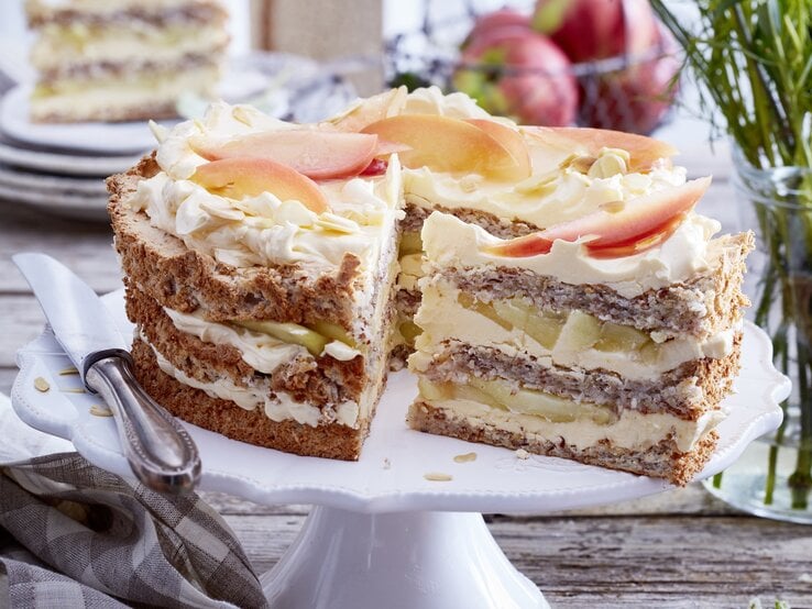 Schwedische Apfel-Mandeltorte angeschnitten auf einem weißen Tortenteller, darauf liegt ein Messer und links davon liegt ein Geschirrtuch. Der Hintergrund ist hölzern und hinten auf dem Foto eine Schüssel mit Äpfeln und kleine Teller gestapelt - darauf ein Stück Torte.