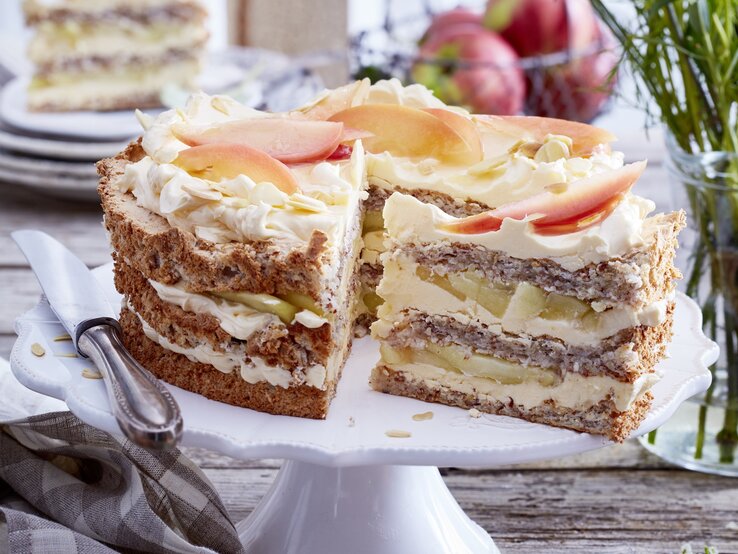 Schwedische-Apfel-Mandeltorte Schwedische Apfel-Mandeltorte angeschnitten auf einem weißen Tortenteller, darauf liegt ein Messer und links davon liegt ein Geschirrtuch. Der Hintergrund ist hölzern und hinten auf dem Foto eine Schüssel mit Äpfeln und kleine Teller gestapelt - darauf ein Stück Torte.