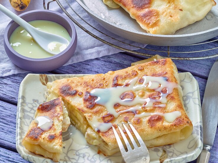 Der Bananen-Pfannenstrudel auf einem eckigen hellen Teller mit heller Soße. Alles auf einem blau-violetten Untergrund.