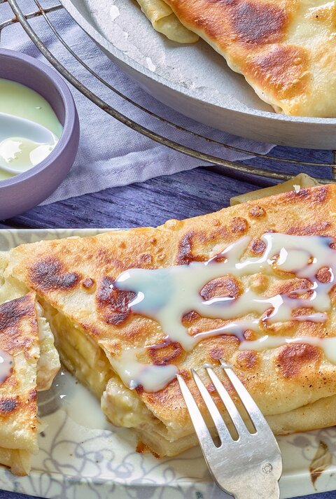 Der Bananen-Pfannenstrudel auf einem eckigen hellen Teller mit heller Soße. Alles auf einem blau-violetten Untergrund.