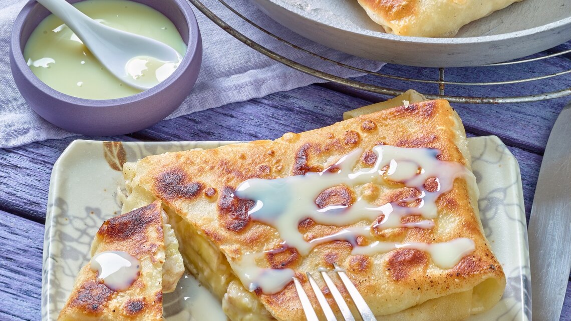 Der Bananen-Pfannenstrudel auf einem eckigen hellen Teller mit heller Soße. Alles auf einem blau-violetten Untergrund.