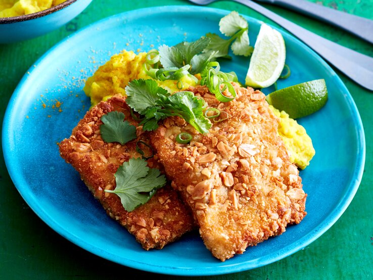 Erdnuss-Schnitzel mit Curry-Stampf auf einem Teller serviert. Rechts davon Besteck. Draufsicht.