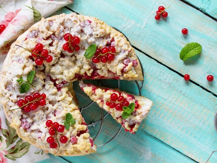 Johannisbeerkuchen mit Streusel auf einem Kuchengitter vor blauem Hintergrund. Links eine Tischdecke.