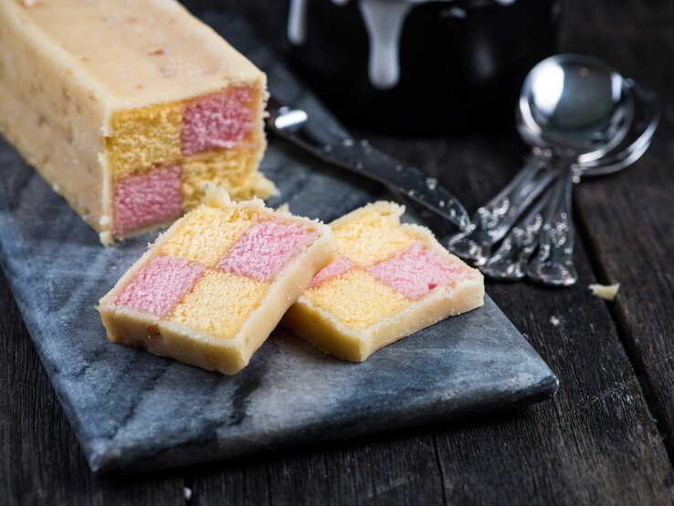 Angeschnittener britischer Battenburg-Kuchen auf einer dunklen Steinplatte auf einem dunklen Holztisch.