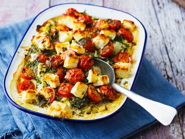 Eine weiße Auflaufform mit blauem Rand und dem Gericht auf einem rustikalem Holztisch mit blauer Tischdecke fotografiert