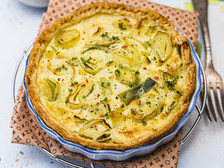 Eine goldbraun gebackene Quiche mit Lauchscheiben in einer runden Backform mit blauem Rand.
