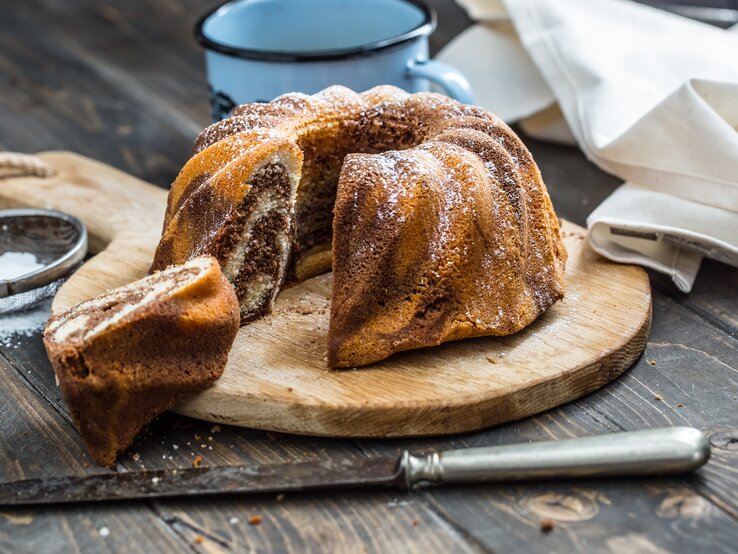 Ein frisch gebackener Marmorkuchen in Gugelhupfform, bestäubt mit Puderzucker, präsentiert auf einem Holzbrett. 