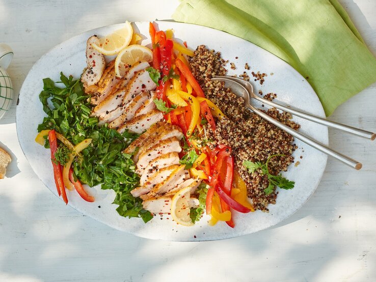 Eine kunterbunte Leckerei ist unser Quinoa-Hähnchen-Salat mit Rucola auf jeden Fall. Quinoa-Hähnchen-Salat mit Rucola auf einem weißen Teller vor hellem Hintergrund, darauf ein grünes Tuch.