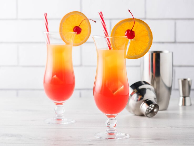 Zwei mit einer Orangenscheibe dekortiere Cocktails mit einem Farbverlauf von Rot zu Gelb, was an einen Sonnenaufgang erinnert. 