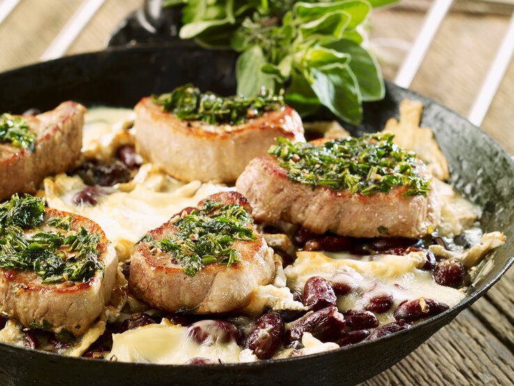 Schweinefilets mit Kräuterkruste auf Kidneybohnen mit Käse überbacken. Alles in der hohen Pfanne auf einem Holztisch fotografiert.