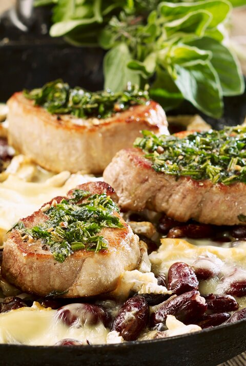 Schweinefilets mit Kräuterkruste auf Kidneybohnen mit Käse überbacken. Alles in der hohen Pfanne auf einem Holztisch fotografiert.
