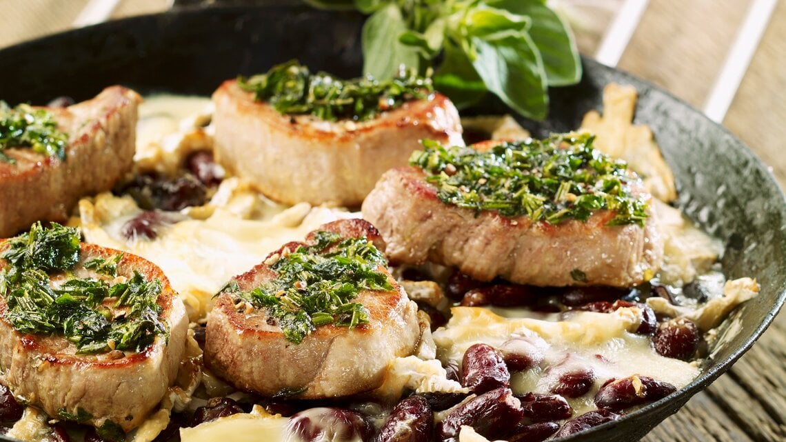 Schweinefilets mit Kräuterkruste auf Kidneybohnen mit Käse überbacken. Alles in der hohen Pfanne auf einem Holztisch fotografiert.