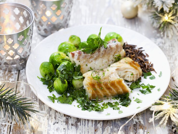Kabeljaufilet mit Rosenkohl Ein festlich dekoriertes Tellergericht mit gebratenem Fisch, frischen Rosenkohl und Wildreis, garniert mit frischen Kräutern, auf einem rustikalen Holztisch. Im Hintergrund sind weihnachtliche Dekorationen wie Zweige und Kerzen zu sehen.